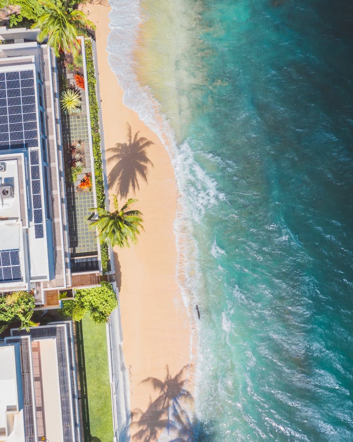 Aerial shot of a solar-powered building by a tropical beach with lush greenery and crashing waves.